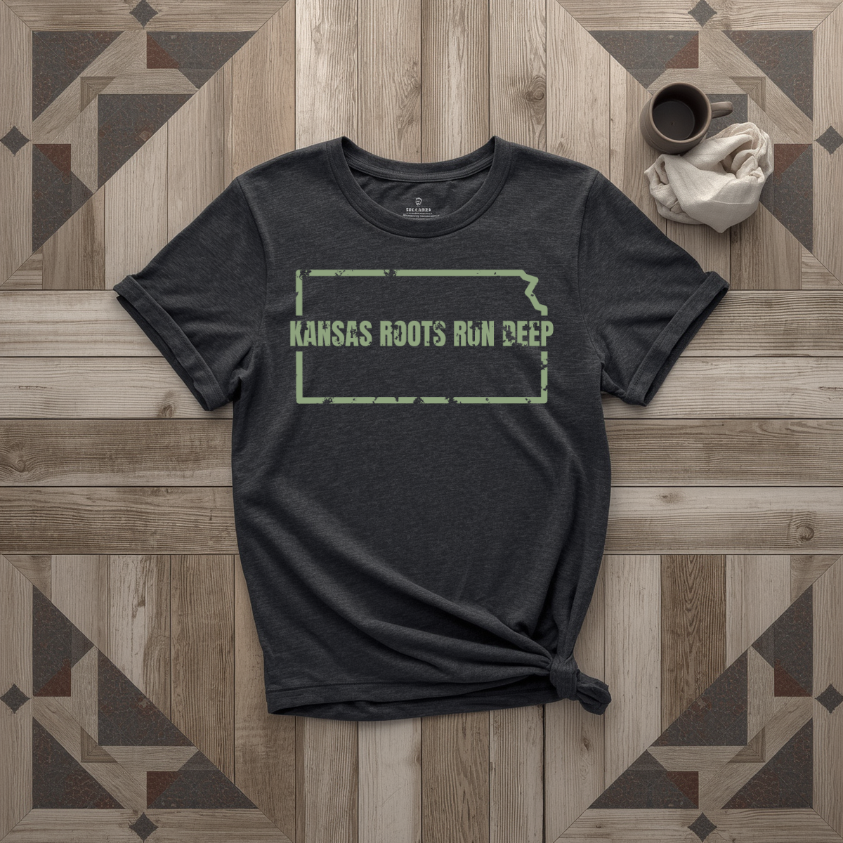 Kansas Roots Run Deep tee in black with sage print, laid flat on weathered wood