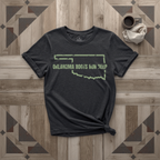 Oklahoma Roots Run Deep tee in black with sage print, laid flat on weathered wood