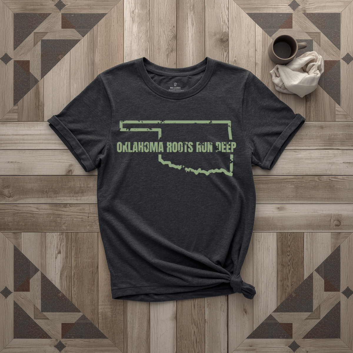 Oklahoma Roots Run Deep tee in black with sage print, laid flat on weathered wood