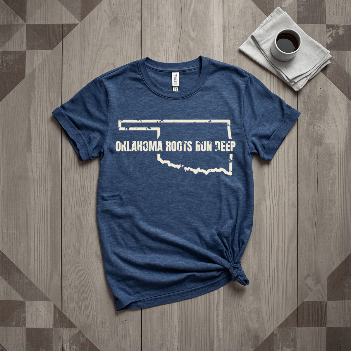 Oklahoma Roots Run Deep tee in heather navy with cream print, laid flat on weathered wood